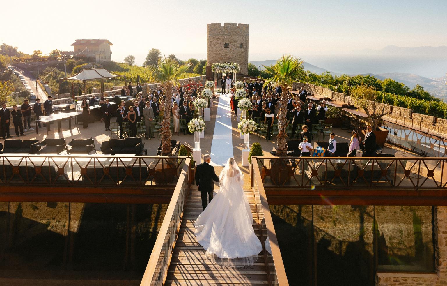 Newlyweds walk down the catwalk towards an outdoor wedding ceremony in a panoramic castle, surrounded by guests and elegant floral arrangements.