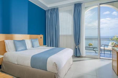 Hotelzimmer mit Meerblick mit Doppelbett, hellblauen Möbeln und Panoramabalkon mit Blick auf das Meer.
