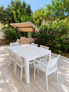 Arredo da giardino bianco con tavolo e sedie, dondolo e piante verdi su terrazza esterna.