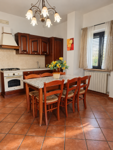 Cucina rustica in legno scuro con grande tavolo da pranzo, sedie in legno, piano piastrellato e fiori gialli al centro.